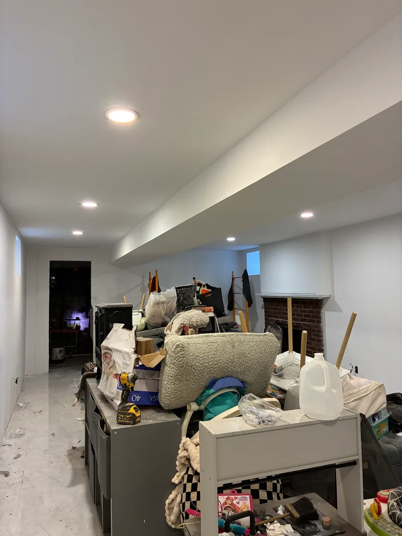 Basement during renovation with new white drywall, recessed ceiling lights, a brick fireplace surround, and stored furnishings in the work area.