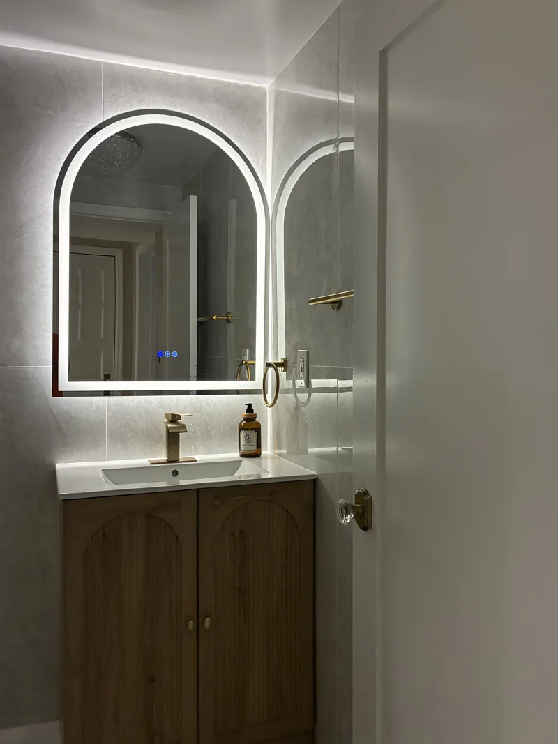 Bathroom with a wood vanity, an arched backlit mirror, a brass faucet, and large-format tile walls.