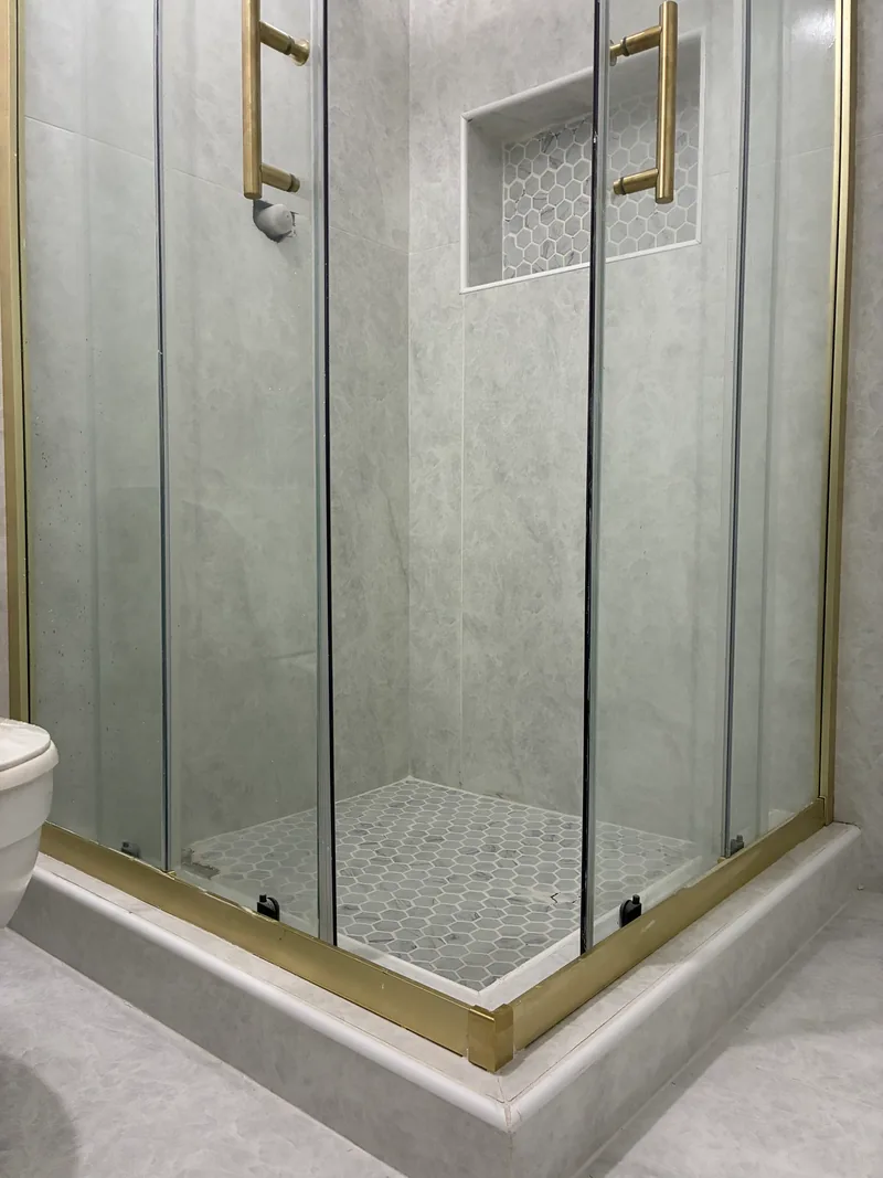 Glass-enclosed shower with marble-look tile, brass frame and handles, a recessed mosaic niche, and a hexagonal tile floor in a renovated bathroom.
