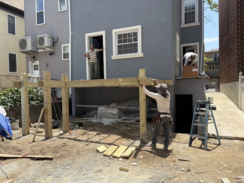Rear deck framing with new lumber, with workers setting posts against a gray-sided house.