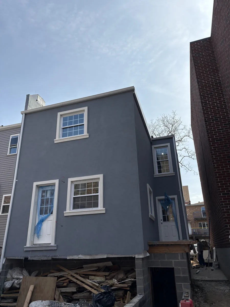 Rear elevation with new gray stucco, replacement windows with white trim, and construction materials below during exterior work.