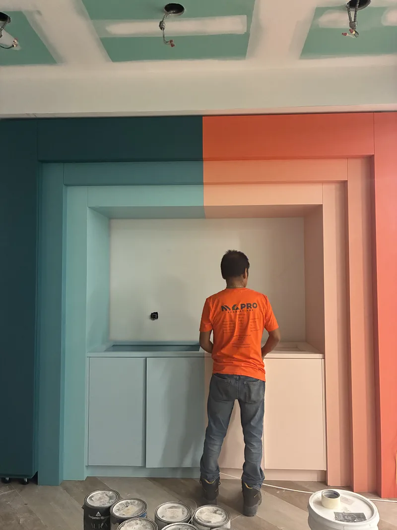 Built-in alcove with teal and coral painted walls, white cabinetry, and a worker applying finish paint.