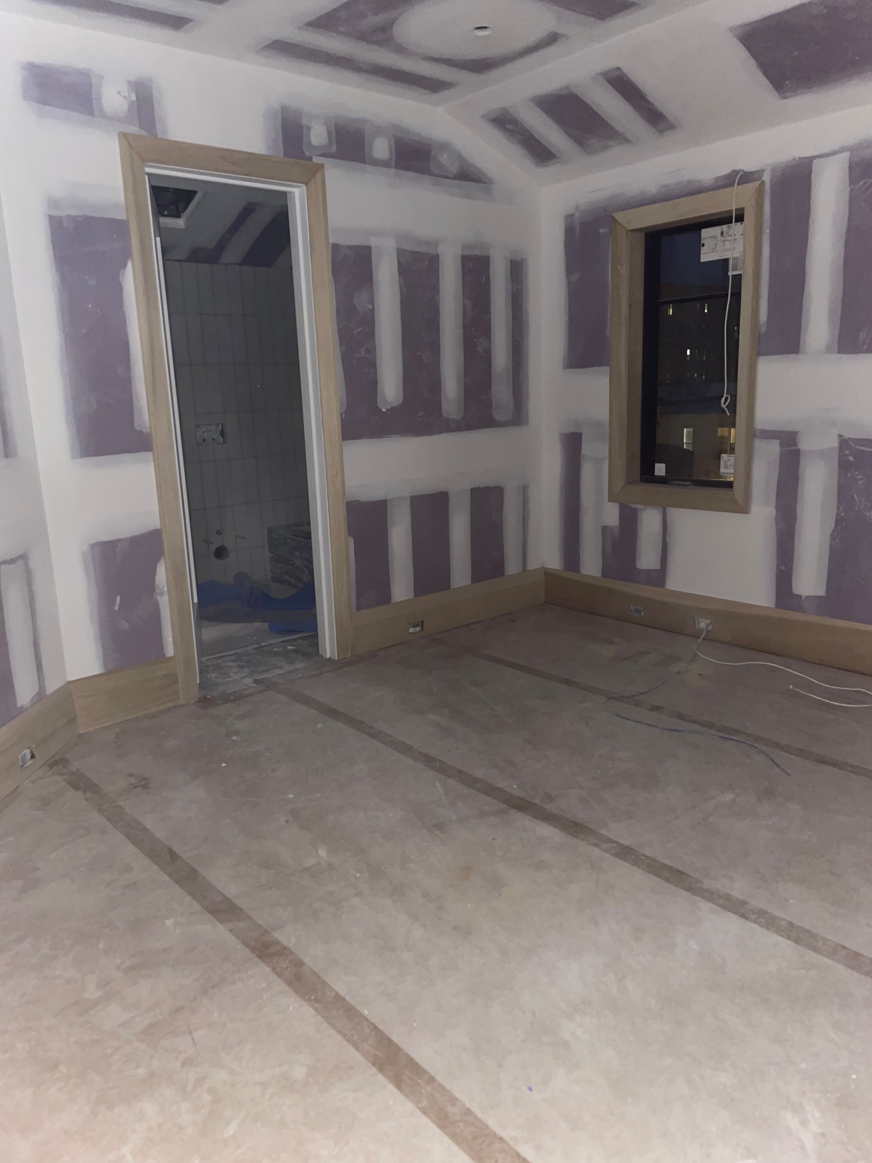 Interior room during renovation, with patched drywall, unfinished wood door and window trim, and a large-format tile floor.